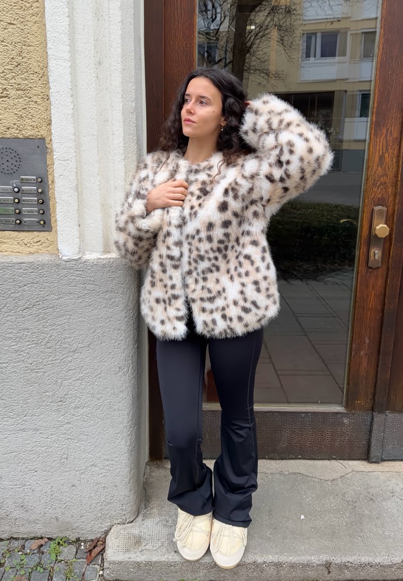 Veste en fausse fourrure à motifs léopard, tons crème et marron, coupe ample ; associée à un pantalon noir ajusté et des chaussures crème.