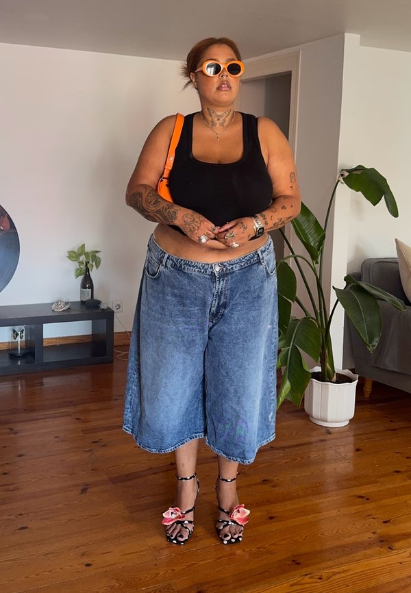 Persoon met tatoeages die een zwart tanktopje, oversized denim shorts, oranje zonnebril en hakken met roze bloemen draagt, staande binnen.