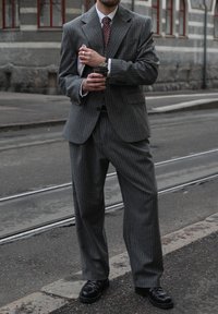 Grijs krijtstreep pak met een notched revers, wit overhemd en rood gestreepte das. Zwarte schoenen. Houdt een mok en een notitieboekje vast. Buitenomgeving.