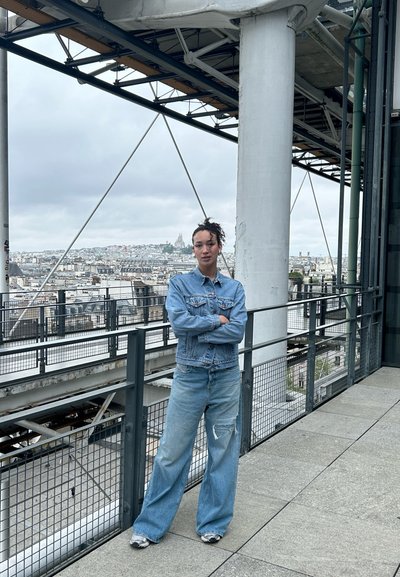 Chaqueta de mezclilla y jeans de pierna ancha en azul claro, con detalles desgastados; de pie contra una barandilla de metal con vista a la ciudad.