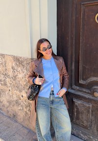 Femme souriante appuyée contre un mur de pierre et une porte en bois, portant des lunettes de soleil, un cardigan bleu, une veste en cuir marron et un jean ample.