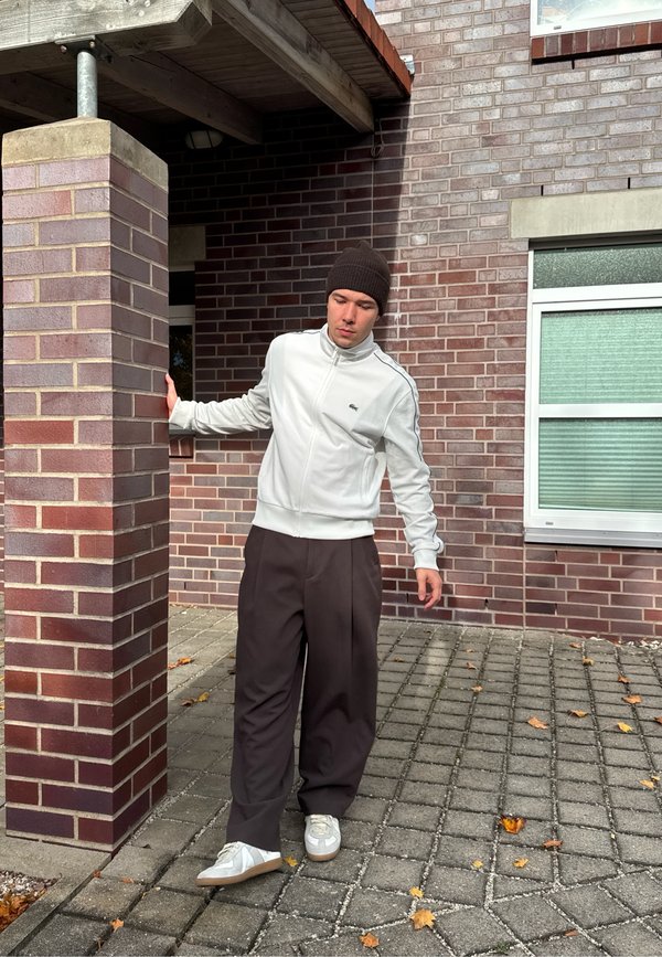A young man dressed in a white jacket, black trousers, and a beanie is stepping outside, holding onto a brick pillar, with a brick building and a window visible in the background.