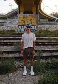 Sportos férfi alak áll a gravell mellett a vasútvonalnál, fehér pólót, szürke sortot és fehér zoknit viselve, sportcipőben.