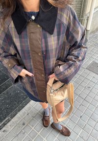 Persona, kas ģērbta raibā jaku ar lielu melnu apkakli, brūnām mokasīnām, pelēkām zeķēm, tur nelielu bēškrāsas rokassomu uz flīzēta ietves.