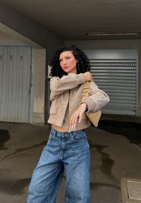 Beige kort jakke med myk tekstur, båret over en matchende topp og sammen med løse blå denimjeans. Flettet beige veske og sølvring.