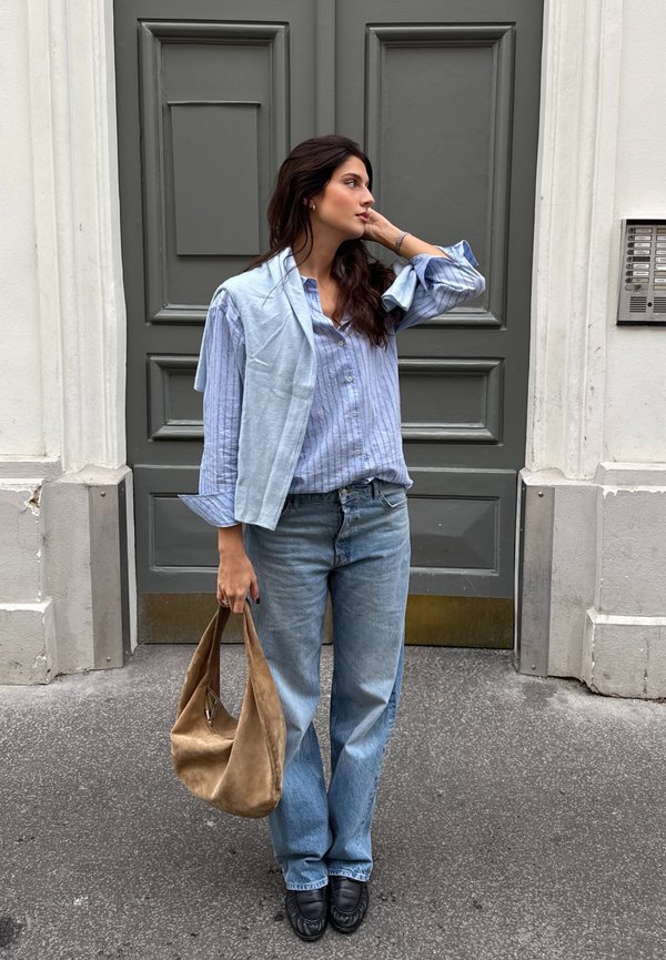 Chemise rayée bleu clair avec les manches retroussées, assortie à un jean bleu clair. Tenant un sac en suede beige, debout devant une porte grise.