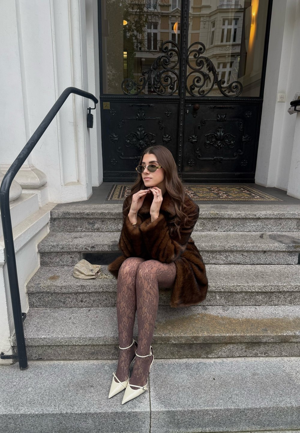 Ruda kailinė striukė, permatomos nėriniuotos pėdkelnės, balti smailūs aukštakulniai, akiniai nuo saulės, sėdinti ant akmeninių laiptų, juodos puošnios durys fone.