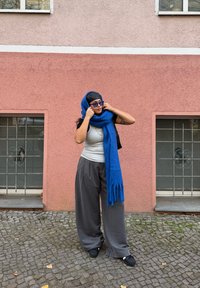 Personne portant un débardeur gris et un pantalon large gris, drapée dans une longue écharpe bleue. L'arrière-plan est un mur rose avec deux fenêtres.