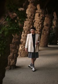 Model nosí krémový blazer, čierne tričko a šedé šortky, so bielymi topánkami a obdĺžnikovými slnečnými okuliarmi, kráčajúc v prostredí kamenného oblúka.
