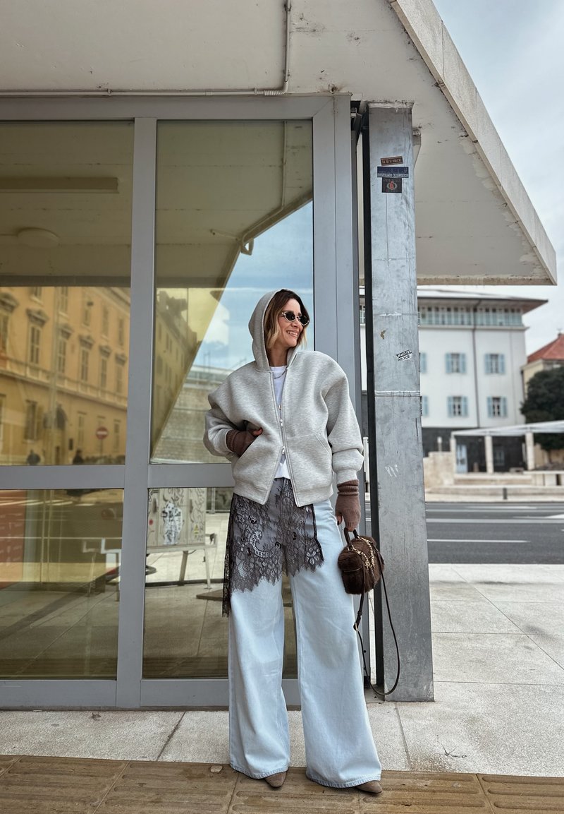 Femme portant un sweat à capuche gris, pantalon large avec bordure en dentelle, lunettes de soleil et gants, debout près de portes en verre dans un cadre urbain.
