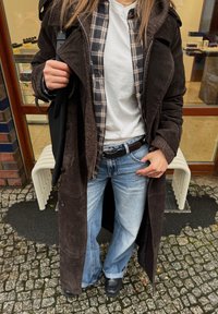 Hnedý semišový dlhý kabát cez károvanú košeľu a biele tričko, v kombinácii s svetlomodrými rozšírenými džínami a čiernymi topánkami. Čierna taška cez rameno.