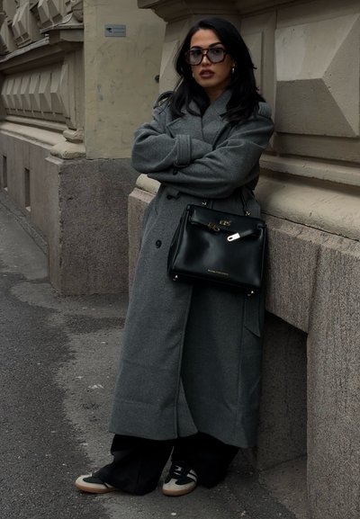 Abrigo gris de doble botonadura, bolso de cuero negro con acentos dorados y pantalones anchos negros. Zapatos oscuros con detalles en blanco y verde.