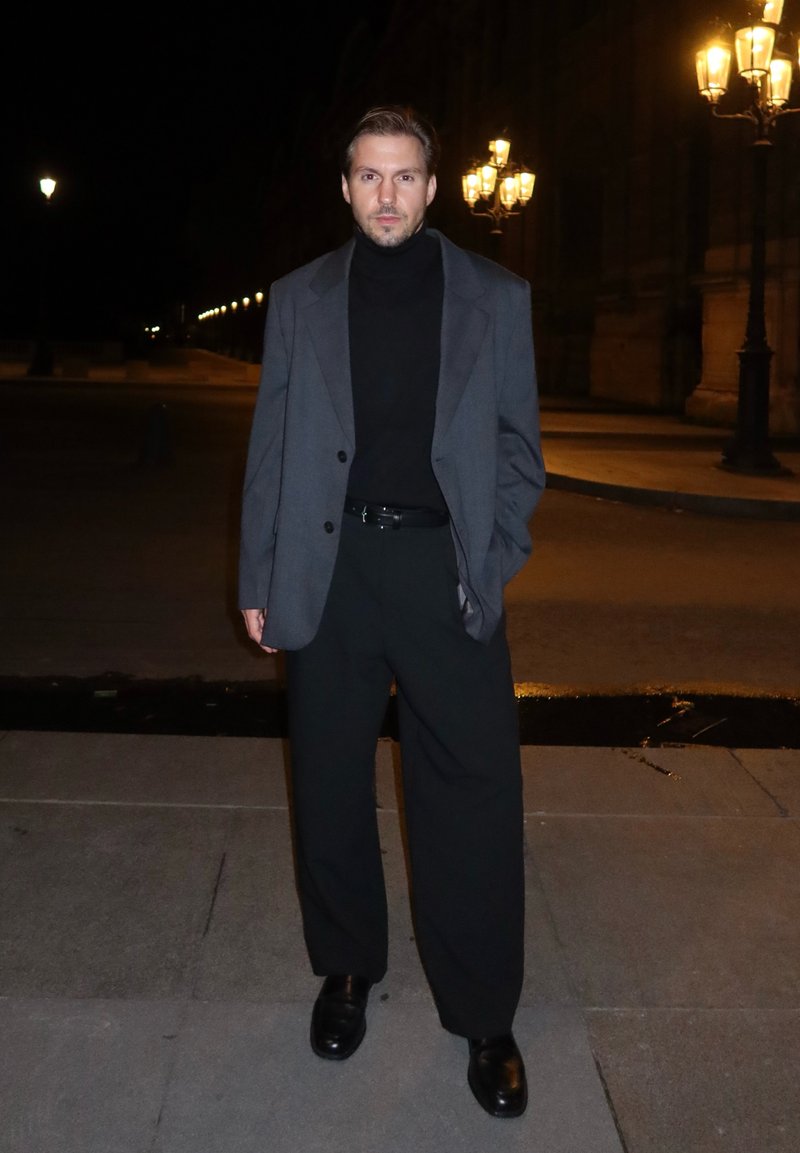 Maglione nero a collo alto, blazer grigio oversize e pantaloni neri a gamba larga. Scarpe nere lucide. I lampioni illuminano lo sfondo notturno.