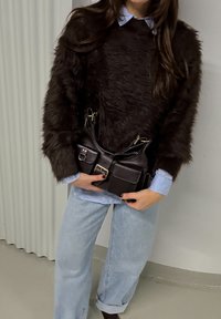 Personne portant un pull en fourrure marron foncé par-dessus une chemise bleu clair, un jean bleu clair, tenant un sac à main en cuir noir avec des poches frontales et une boucle.