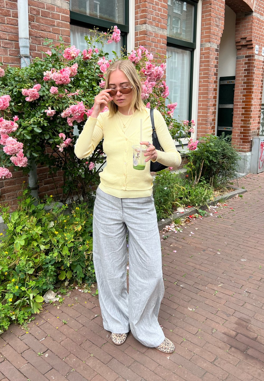 Hellgelbes geribbeltes Cardigan mit Knöpfen, graue weit geschnittene Leinenhose und Schuhe im Leopardenmuster. Hält ein klares Getränk vor blühenden Büschen.