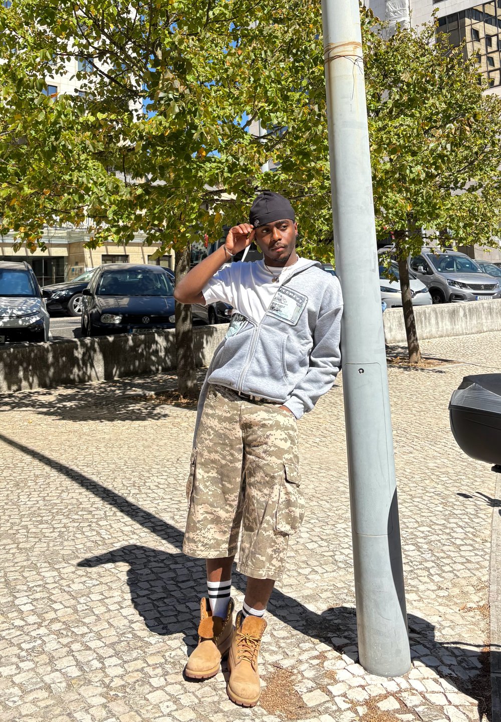 Hanorac gri cu fermoar și un design grafic, pantaloni scurți cargo cu model camuflat și cizme bej. Stă lângă un stâlp de lumină într-o zonă exterioară pavate.