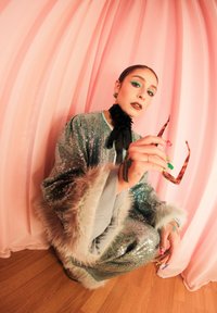 Tenue turquoise scintillante avec accents en fourrure, nœud papillon noir et lunettes de soleil à motifs. Fond rose pâle et sol en bois mettent en valeur le décor.