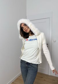Ekrü színű cipzáras kardigán bolyhos, fehér kapucnival, alatta egy világos póló kék "I ♥ CRYING" felirattal, sötét farmernadrággal párosítva.