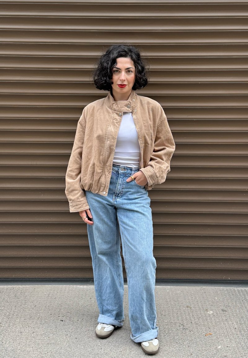 Tan kordjacka i manchester, vit tät topp, högmidjade ljusblå jeans och beige skor med grå accenter. Bakgrunden har horisontella linjer.