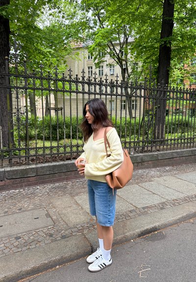 Suéter amarillo claro, pantalones cortos de mezclilla azules y calcetines blancos combinados con zapatillas Adidas. Bolso de cuero marrón sostenido con una bebida fría.