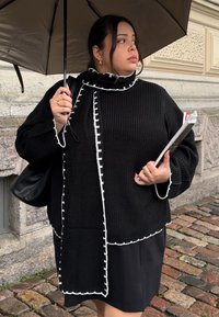 Melns adīts džemperis ar baltu apmali, ar brīvu piegriezumu, augstu apkakli un lielām piedurknēm. Valdīts ar melnu svārku un tur lietussargu.