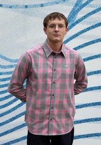 Hall ja roosa ruuduline särk, millel on nööbid, pikad varrukad, terav krae ja väikese rinnamärgiga, kombineeritud tumedate teksadega.