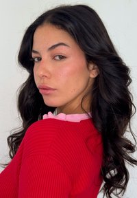 Rød ribstrikket sweater med en pink flæsekrave; langt mørkt hår med bløde krøller, placeret mod en neutral baggrund.