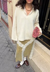 Femme portant un pull oversize crème, une jupe midi olive ornée de dentelle, des mocassins marron, des chaussettes blanches, un collier chaîne en or, tenant une pochette rose plissée.