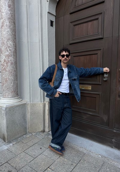 Chaqueta de mezclilla y jeans oscuros de corte holgado, combinados con una camiseta blanca. Accesorios incluyen gafas de sol y un bolso de hombro marrón.