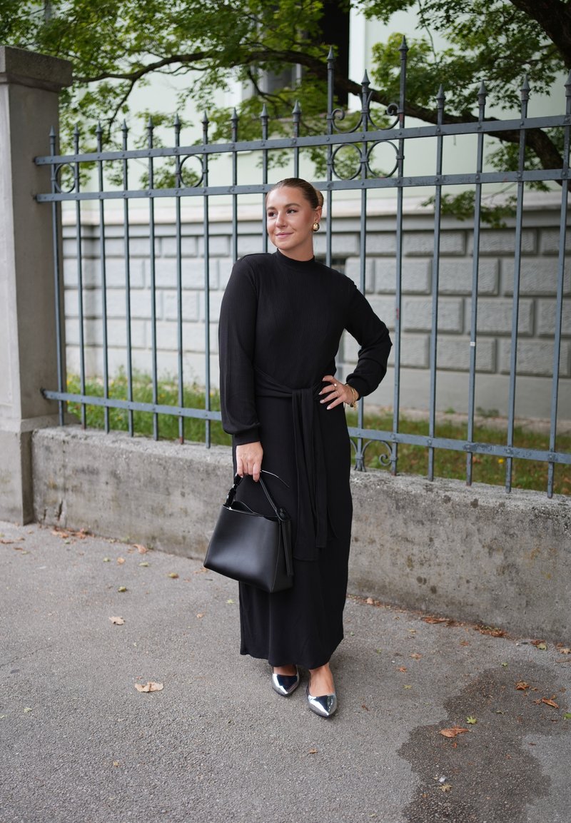 Robe noire à manches longues côtelées avec une taille à nouer, associée à un sac à main noir et des ballerines argentées à bouts pointus, sur fond de portail.