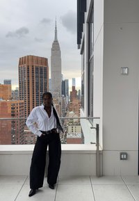 Witte overhemd met knopen, zwarte broek met wijde pijpen en zwarte enkellaarzen. Stadsgezichten met het Empire State Building op de achtergrond. Glazen balustrade.