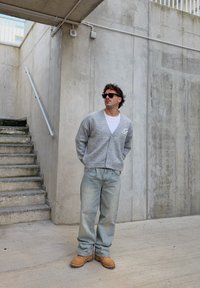 Cardigan grigio lavorato a maglia con bottoni, indossato sopra una camicia bianca. Jeans larghi azzurri e stivali da lavoro color cuoio. Sullo sfondo, pareti di cemento e scale.