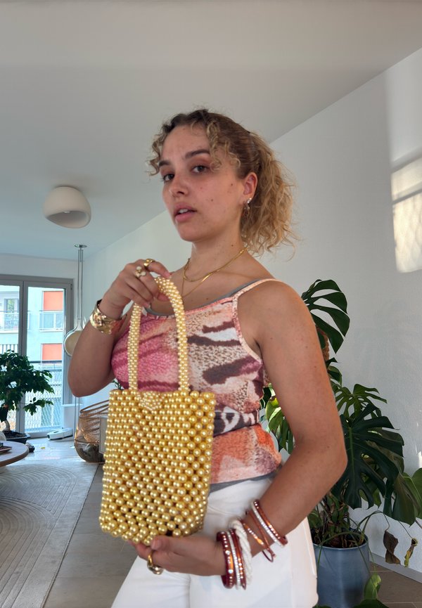 Jeune femme à l'intérieur tenant un sac à main doré orné de perles, portant un haut rose à motifs et un pantalon blanc avec plusieurs bracelets à son poignet.