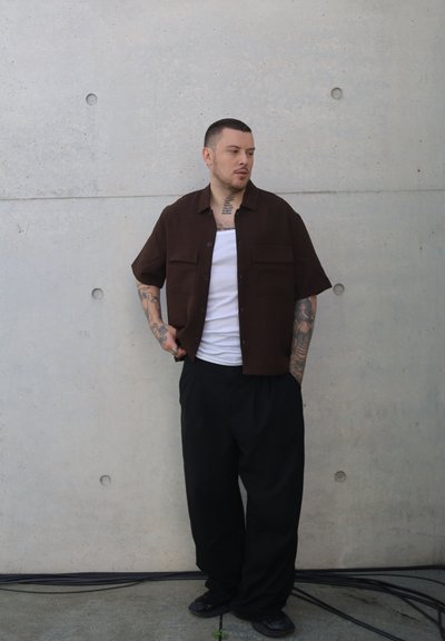 Camisa marrón de manga corta con dos bolsillos de parche, combinada con pantalones negros holgados y una camiseta sin mangas blanca, de pie contra una pared gris.