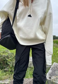 Felpa con zip crema e logo nero, abbinata a pantaloni cargo neri larghi. Una borsa a tracolla nera è indossata su un lato. Sfondo verde.