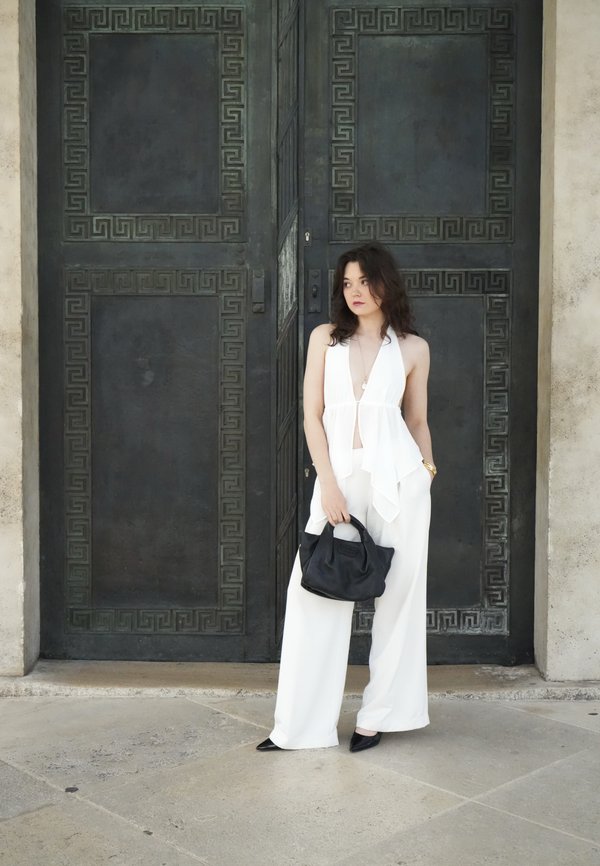 Woman in white sleeveless top and pants holding black bag stands against large dark ornate metal door.