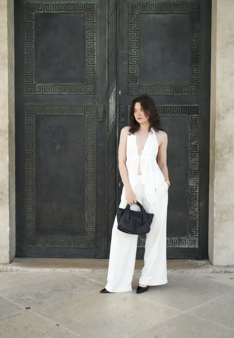 Femme en top blanc sans manches et pantalon tenant un sac noir, debout devant une grande porte en métal sombre ornée.