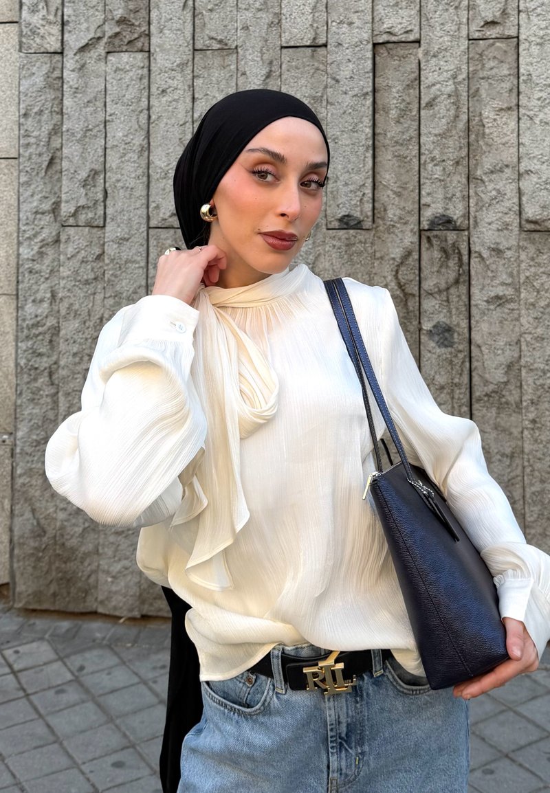 Kobieta w czarnej chustce na głowie i białej bluzce pozuje na tle kamiennego muru, niesie czarną torebkę i nosi niebieskie dżinsy z czarnym paskiem.