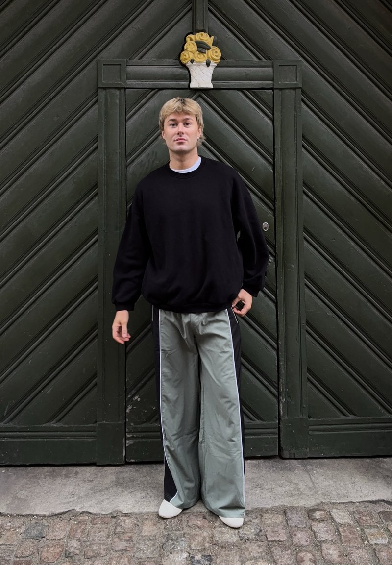 Personne blonde vêtue d'un pull noir et d'un pantalon gris à jambes larges se tient devant une grande porte en bois vert foncé avec une décoration florale jaune.