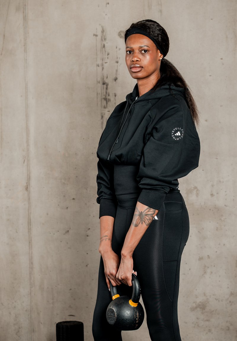 Femme en tenue de sport noire tenant une kettlebell à deux mains devant un mur en béton simple.