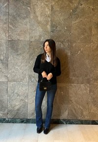 Schwarzer V-Ausschnittpullover, weißes Hemd mit Kragen, weit ausgestellte blaue Jeans, schwarze Handtasche mit strukturiertem Finish und schwarze Schuhe vor einem Marmorhintergrund.