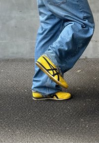 Geltoni sportiniai bateliai su juodais akcentais, margintu dizainu ir tekstūruotais padą. Asmuo vilki išblukusius mėlynus džinsus su laisvu kirpimu.
