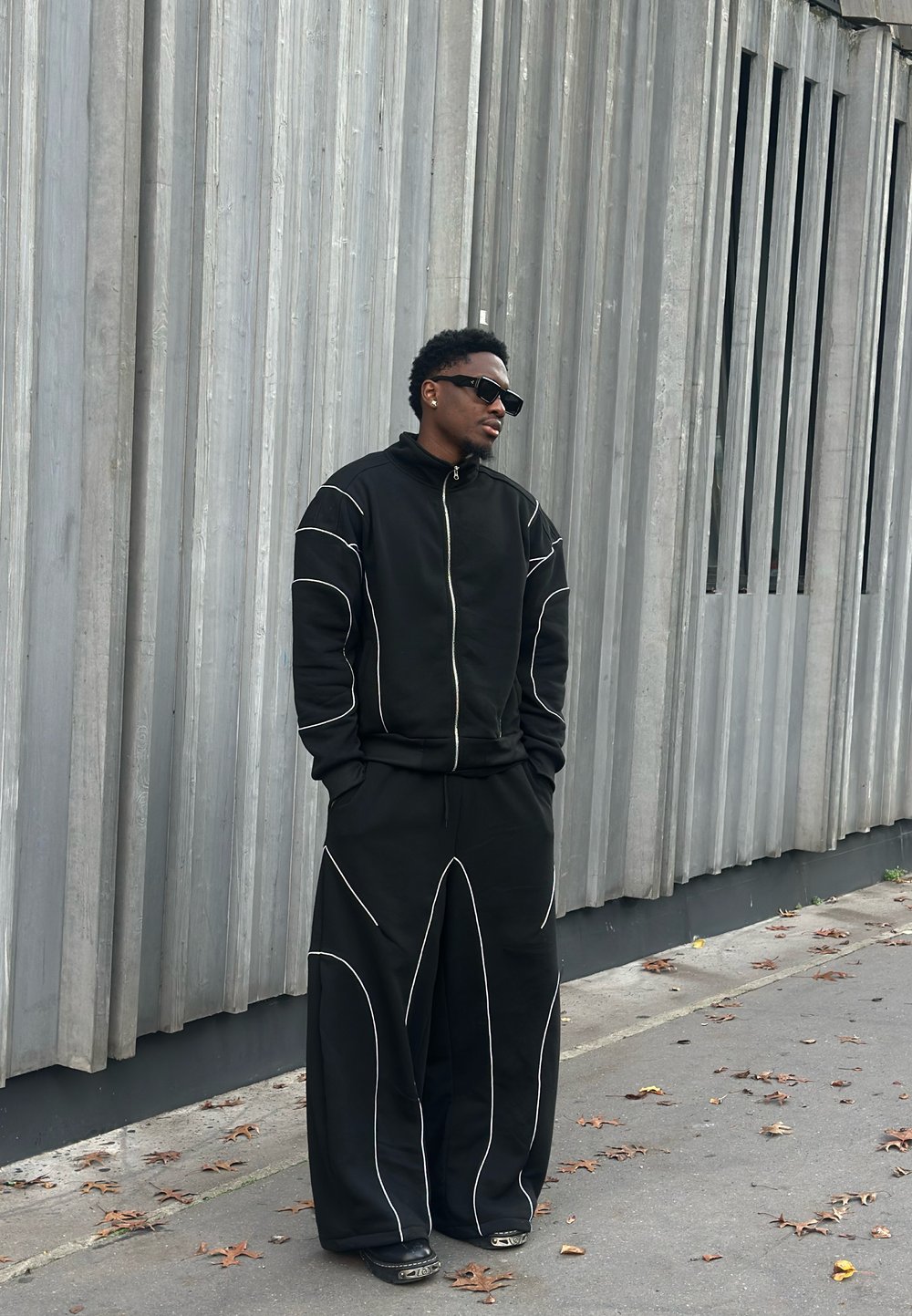 Chaqueta negra con cremallera y pantalones anchos con detalles de costuras blancas. Llevados con gafas de sol; sobre un fondo gris con paneles verticales.