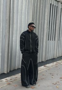 Chaqueta negra con cremallera y pantalones anchos con detalles de costuras blancas. Llevados con gafas de sol; sobre un fondo gris con paneles verticales.