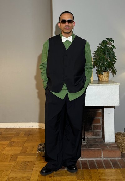 Hombre con gafas de sol, pajarita blanca, camisa verde, chaleco negro y pantalones anchos negros, de pie en el interior junto a una planta sobre una repisa.