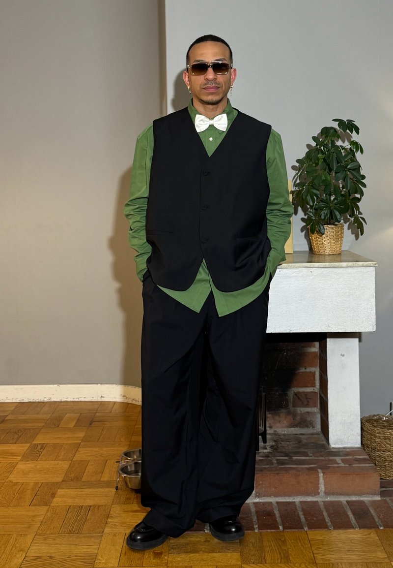 Homme portant des lunettes de soleil, un nœud papillon blanc, une chemise verte, un gilet noir et un pantalon large noir, debout à l'intérieur à côté d'une plante sur une cheminée.