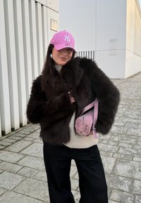 Femme portant une casquette New York Yankees rose, un manteau en fausse fourrure noire, un pull beige, un pantalon noir, et portant un petit sac rose sur un trottoir en ville.