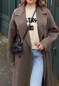 Femme portant un manteau en laine marron, un pull crème avec un texte noir, un jean bleu clair et un petit sac à main en cuir noir avec une bandoulière traversant le corps.