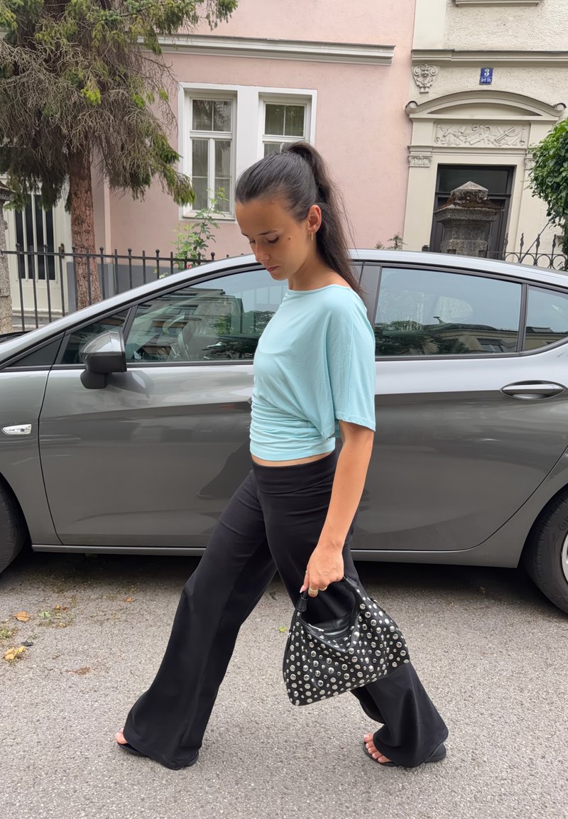 Femme en haut bleu clair et pantalon noir marchant dans la rue, tenant un sac à main noir clouté, une voiture grise et des bâtiments en arrière-plan.