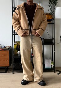 Veste zippée en shearling marron avec coutures contrastantes, accents noirs, associée à un jean large beige clair et des bottes noires épaisses.